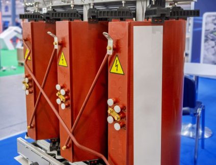 A close-up view of a modern industrial dry-type transformer, featuring three large, reddish-brown coils prominently displaying yellow high voltage warning symbols. The intricate design includes multiple copper connections, insulators, and robust metallic structural components, indicating its role in high-power electrical distribution. This equipment is mounted on a sturdy frame with rollers, suggesting its potential for mobility within an industrial setting or an exhibition. The blurred background hints at a modern factory floor, power plant, or technology trade show, emphasizing its relevance in contemporary electrical engineering and infrastructure projects. This image is ideal for illustrating topics related to energy transmission, industrial automation, electrical safety, or advanced manufacturing.